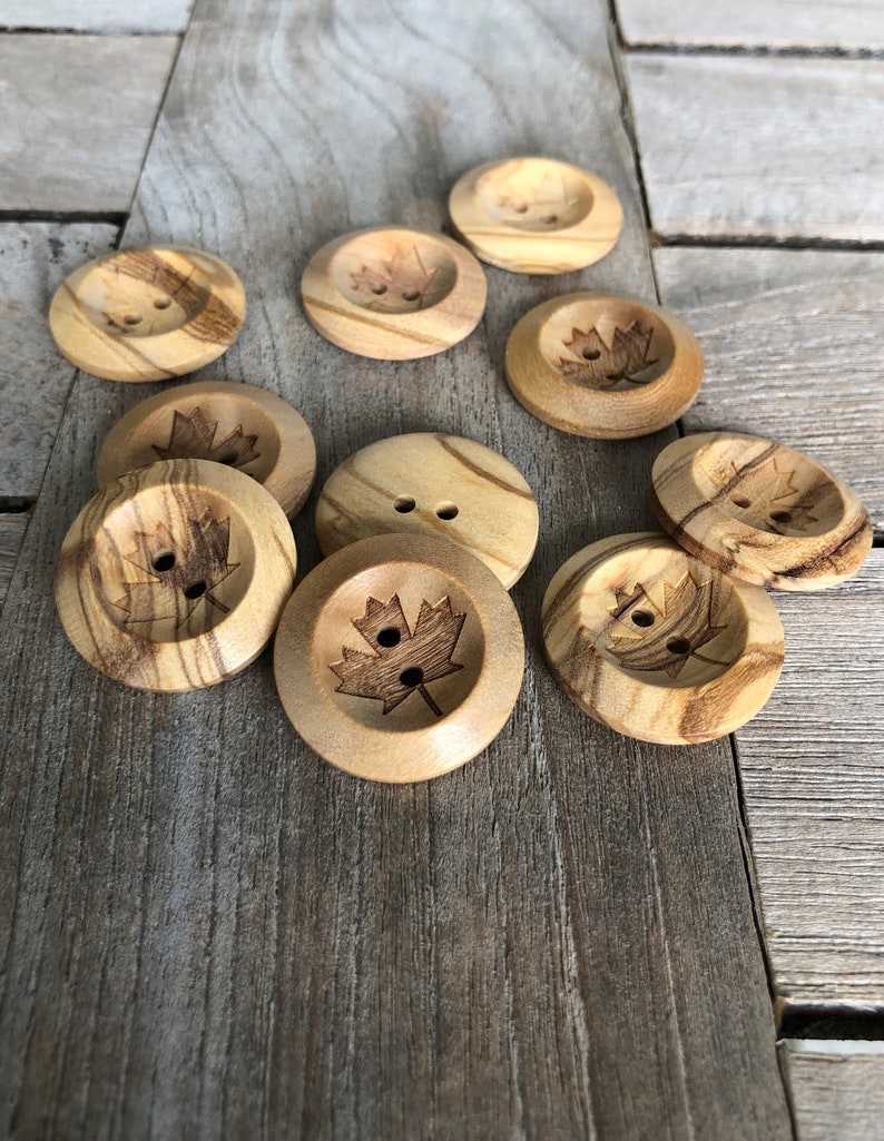 May include: A collection of ten wooden buttons with a maple leaf design. The buttons are a light brown color with darker brown wood grain patterns.