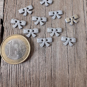 May include: A collection of ten silver glitter bow-shaped buttons on a wooden surface. A Euro coin is next to the buttons for size comparison. The buttons have a textured, sparkly appearance and are evenly spaced.