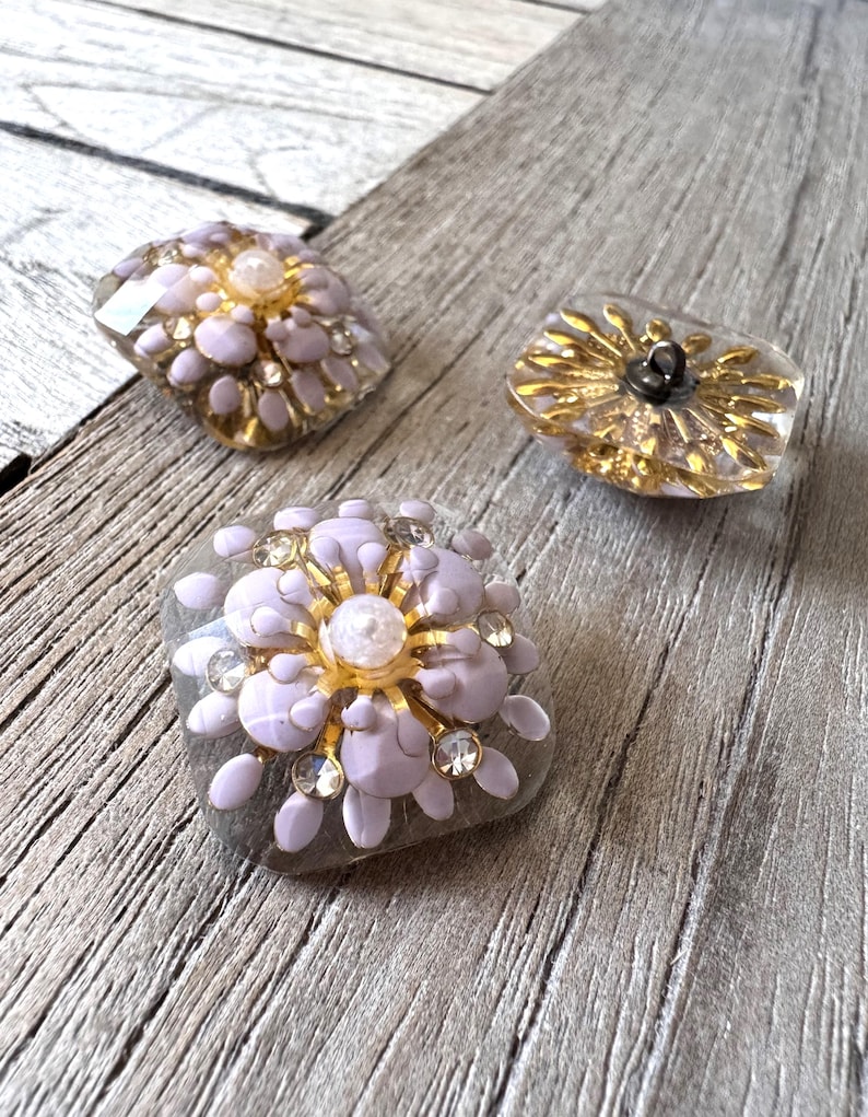 May include: Three decorative buttons with floral designs. The buttons feature a combination of gold, lavender, and clear resin. Each button has a unique design, with some incorporating rhinestones for added sparkle.