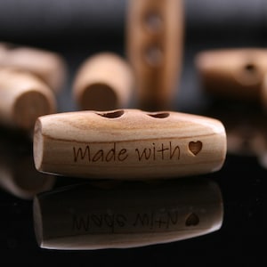 May include: Close-up of several wooden toggle buttons. The buttons are light brown with a smooth finish. One button is in focus, displaying the words "Made with" and a heart symbol. The buttons are likely for sewing or crafts.