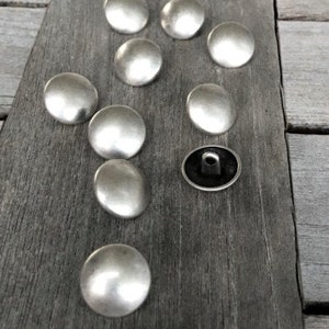 May include: A collection of ten silver-toned metal buttons, each with a rounded, slightly domed shape. The buttons are arranged on a wooden surface.