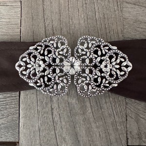 Könnte beinhalten: Silberne filigranartige Gürtelschnalle mit floralem Design. Die Schnalle ist aus Metall und hat ein zartes, filigranes Muster.