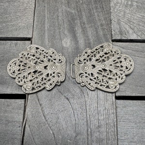 K&ouml;nnte beinhalten: Ein Paar silberne Filigran-Metallschlie&szlig;en mit komplizierten Designs. Die Schlie&szlig;en sind symmetrisch und haben eine zarte, verzierte Optik.