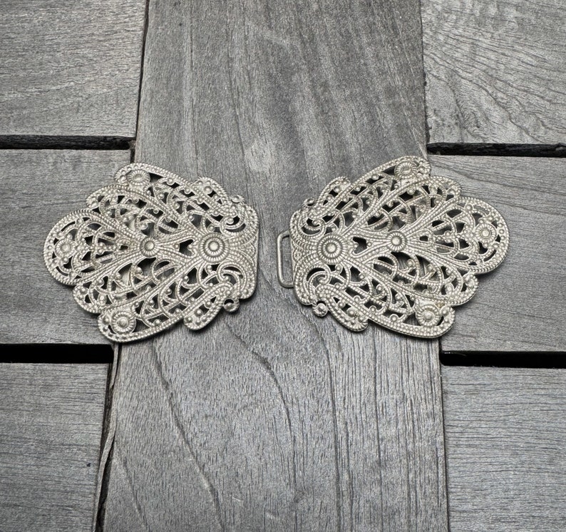 K&ouml;nnte beinhalten: Ein Paar silberne Filigran-Metallschlie&szlig;en mit komplizierten Designs. Die Schlie&szlig;en sind symmetrisch und haben eine zarte, verzierte Optik.