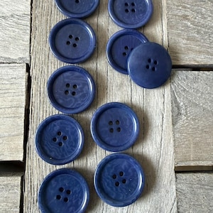 Könnte beinhalten: Eine Ansammlung marineblauer Knöpfe mit jeweils vier Löchern, angeordnet auf einer verwitterten Holzoberfläche. Die Knöpfe sind rund und haben ein leicht marmoriertes Aussehen. Ideal für Näh- und Bastelprojekte.