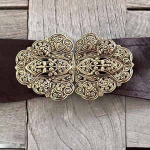 Puede incluir: Una hebilla de cinturón de metal dorado con un diseño filigranado detallado. La hebilla es ornamentada y presenta un patrón simétrico de espirales y bucles.