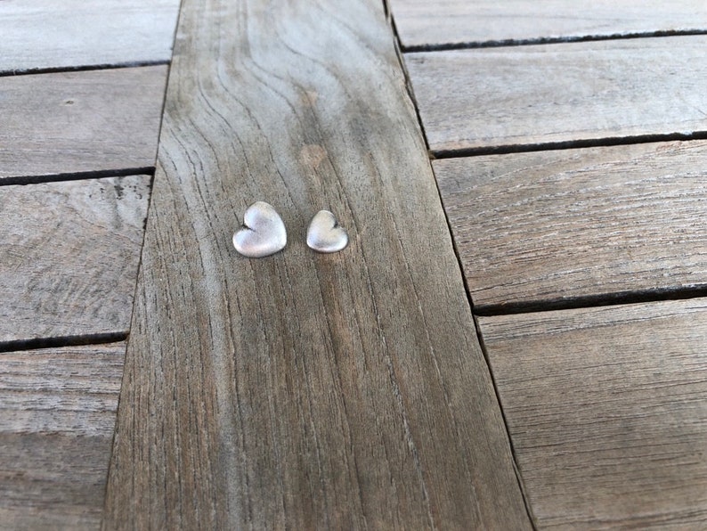 May include: Two silver heart-shaped charms on a wooden surface.