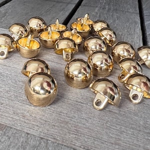 Op de afbeelding: Een verzameling goudkleurige metalen belbedels, sommige met een lus om op te hangen. De bellen zijn in verschillende stadia van montage, sommige met de klepel erin en andere zonder. De bedels zijn verspreid over een houten oppervlak.
