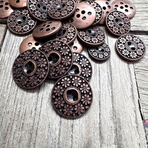 May include: A collection of copper-colored metal buttons in various sizes. Some buttons have a floral design around the edges, while others have a simple hole pattern. The buttons are scattered on a wooden surface.