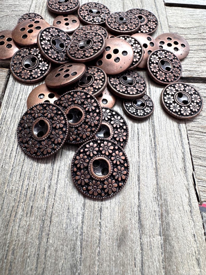 May include: A collection of copper-colored metal buttons in various sizes. Some buttons have a floral design around the edges, while others have a simple hole pattern. The buttons are scattered on a wooden surface.