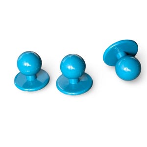 Puede incluir: Tres gemelos de camisa de color azul turquesa. Cada gemelo tiene una parte superior redonda y una base plana y circular. Los gemelos están hechos de un material liso y brillante, sobre un fondo blanco.