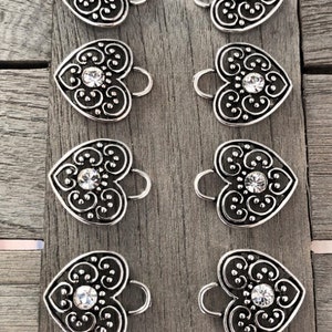May include: Eight silver heart-shaped charms with a clear rhinestone in the center. Each charm has a decorative swirl design and a loop at the top.