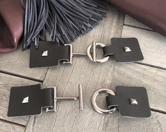 May include: Two black leather belt buckles with silver metal hardware. Each buckle has a square shape with a small pyramid stud in the center. The buckles are connected by a silver metal bar with a ring in the center.
