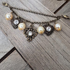 K&ouml;nnte beinhalten: Ein Armband im antiken Stil mit einer Bronzekette und dekorativen Anh&auml;ngern. Das Armband verf&uuml;gt &uuml;ber Kunstperlen und verzierte Filigrananh&auml;nger mit klaren Kristallakzenten. Der Verschluss ist im Karabinerstil. Der Hintergrund ist eine Holzoberfl&auml;che.