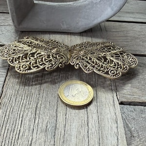 May include: Ornate, antique gold-colored metal clasp with intricate filigree design. The clasp is open, revealing two symmetrical halves. A Euro coin is next to the clasp for size comparison. The clasp is on a weathered wooden surface.