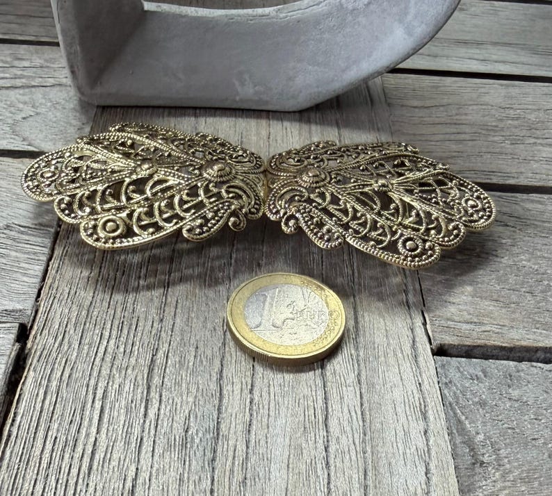 May include: Ornate, antique gold-colored metal clasp with intricate filigree design. The clasp is open, revealing two symmetrical halves. A Euro coin is next to the clasp for size comparison. The clasp is on a weathered wooden surface.