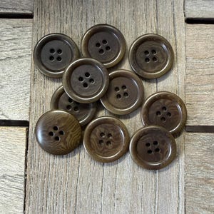Peut inclure: Dix boutons bruns, chacun percé de quatre trous, disposés sur une surface en bois. Les boutons ont un aspect légèrement texturé et une teinte chaude et terreuse. Parfaits pour la couture ou les travaux manuels, environ 2,5 cm de diamètre.