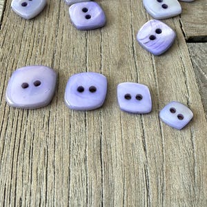 May include: A collection of square, lavender-colored buttons with two holes each, scattered on a wooden surface. The buttons vary slightly in shade and size, with some displaying a marbled effect. Ideal for sewing and craft projects.