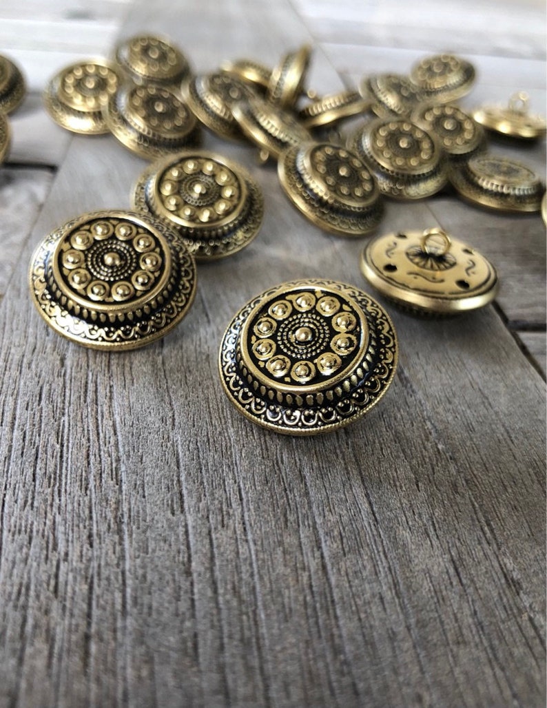 Op de afbeelding: Een groep gouden metalen knopen met ingewikkelde ontwerpen. De knopen hebben een verhoogd midden met een cirkelvormig patroon en een decoratieve rand.