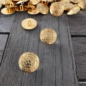 Puede incluir: Un montón de botones de metal de color dorado con un diseño de estrella en el centro. Los botones tienen un borde elevado y un pequeño bucle en la parte posterior. Hay tres botones en primer plano, dos de los cuales tienen un diseño de estrella y uno tiene una superficie lisa.