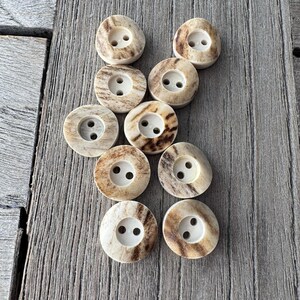 May include: A collection of ten round buttons with two holes each. The buttons have a natural, off-white color with brown marbling. They are arranged on a weathered, gray wooden surface.