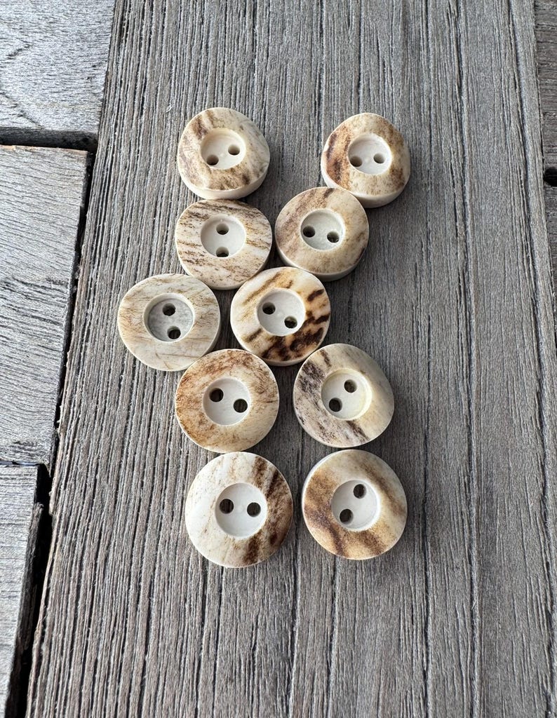 May include: A collection of ten round buttons with two holes each. The buttons have a natural, off-white color with brown marbling. They are arranged on a weathered, gray wooden surface.