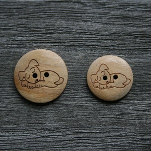 May include: Two wooden buttons with a light brown finish. Each button features a carved illustration of a sleeping dog. The buttons are round, with one slightly larger than the other. The background is a dark gray, textured surface.