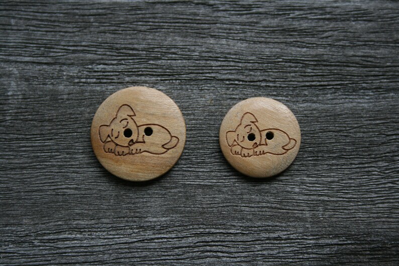 May include: Two wooden buttons with a light brown finish. Each button features a carved illustration of a sleeping dog. The buttons are round, with one slightly larger than the other. The background is a dark gray, textured surface.