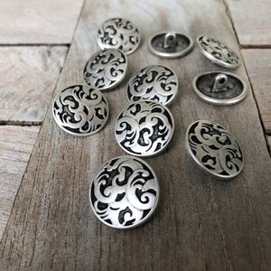 Könnte beinhalten: Ein Satz von zehn silberfarbenen Knöpfen mit einem schwarzen Wirbelmuster. Die Knöpfe sind rund und haben ein erhabenes Design.