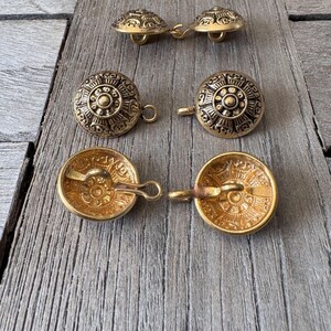 May include: A selection of antique gold-coloured buttons featuring detailed embossed patterns. The buttons are circular with a loop on the reverse for sewing. They are displayed on a weathered wooden surface, highlighting their vintage appeal.