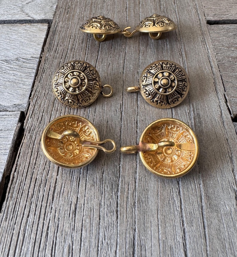 May include: A selection of antique gold-coloured buttons featuring detailed embossed patterns. The buttons are circular with a loop on the reverse for sewing. They are displayed on a weathered wooden surface, highlighting their vintage appeal.
