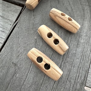 Op de afbeelding: Drie houten toggle-knoppen met elk twee gaten. De knoppen zijn lichtbruin met donkerbruine strepen. Ze zijn in een rij op een houten oppervlak gerangschikt.