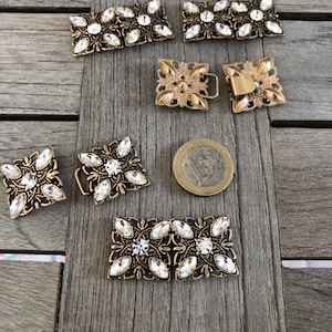 Puede incluir: Un conjunto de ocho hebillas de cinturón de metal dorado antiguo con adornos de diamantes de imitación transparentes. Las hebillas son de varias formas y tamaños, incluyendo cuadradas, rectangulares y ovaladas.