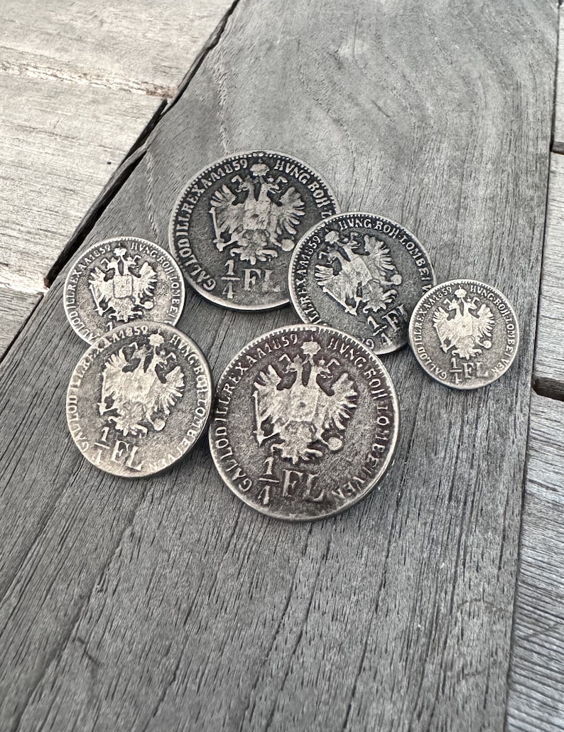 May include: Six silver coins with the inscription "GALLODILUREX A1859 HVNG ROH LOMBEI VEN" and a depiction of a double-headed eagle. The coins are arranged on a wooden surface.