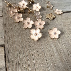 Op de afbeelding: Bloemvormige knopen met een lichtroze emaille afwerking en een klein strass steentje in het midden. De knopen hebben een goudkleurige metalen achterkant en zijn verspreid op een houten oppervlak. Deze decoratieve knopen zijn ideaal voor naai- en knutselprojecten.