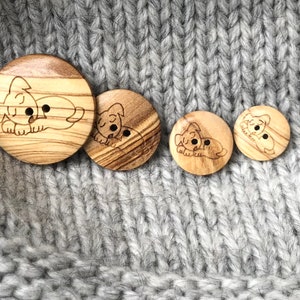 May include: A collection of four wooden buttons with a laser-etched sleeping puppy design. The buttons vary in size and are made of light-colored wood with a visible grain. The buttons are set against a gray knitted fabric background.