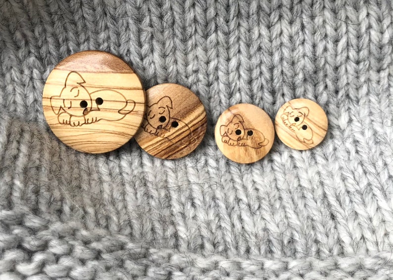 May include: A collection of four wooden buttons with a laser-etched sleeping puppy design. The buttons vary in size and are made of light-colored wood with a visible grain. The buttons are set against a gray knitted fabric background.