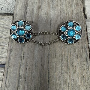 Puede incluir: Dos componentes de broche redondos de color plateado conectados por una cadena. Cada componente presenta un diseño floral con múltiples gemas redondas de color turquesa. La broche está sobre una superficie de madera gris desgastada.