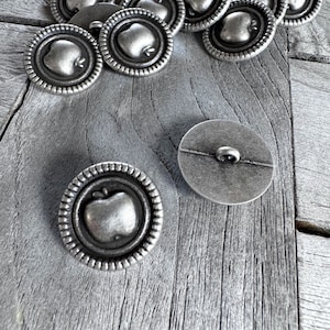 May include: A pile of silver metal buttons with an apple design. The buttons are arranged in a random pattern on a wooden surface.