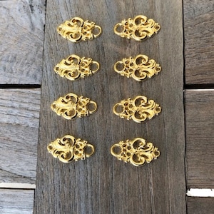 Könnte beinhalten: Acht goldfarbene, metallene Dekorationselemente mit floralem Design. Die Elemente sind oval geformt und haben an jedem Ende ein Loch.