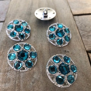May include: Four teal blue rhinestone buttons with silver settings. The buttons have a circular design with a raised center and a decorative border.