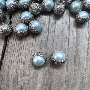 May include: A collection of light blue pearl beads with ornate silver-colored filigree caps. The beads are scattered on a weathered wooden surface, with two beads in the foreground. These beads are suitable for jewelry making and craft projects.