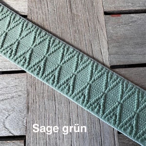 May include: Sage green elastic ribbon with a diamond pattern. The ribbon is displayed on a wooden surface. The text "Sage gr&uuml;n" is visible on the ribbon.