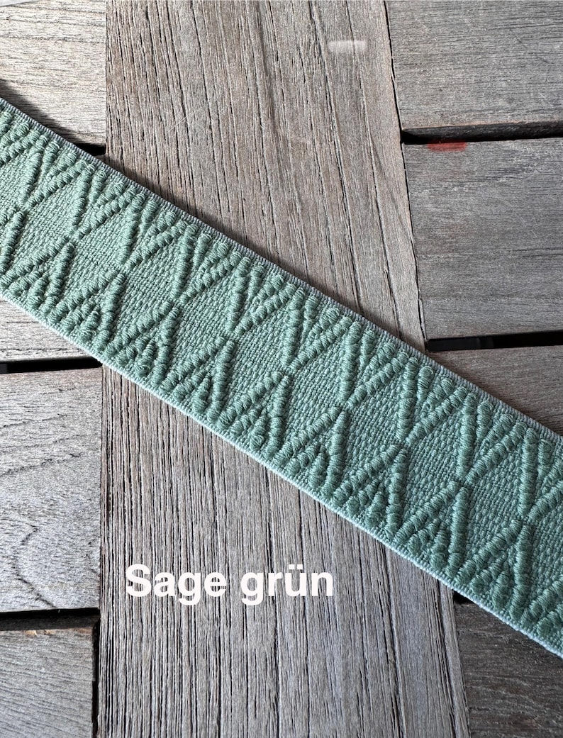 May include: Sage green elastic ribbon with a diamond pattern. The ribbon is displayed on a wooden surface. The text "Sage gr&uuml;n" is visible on the ribbon.