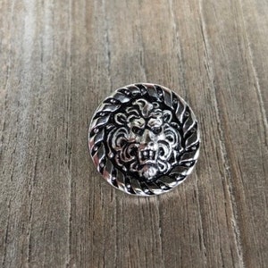 May include: A silver-toned button with a detailed lion head design. The lion's face is surrounded by a black and silver rope-like border.