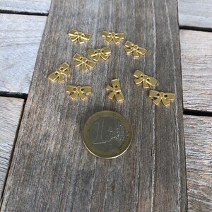 May include: A collection of ten gold-colored metal bow-shaped embellishments scattered on a wooden surface. A euro coin is included for scale.