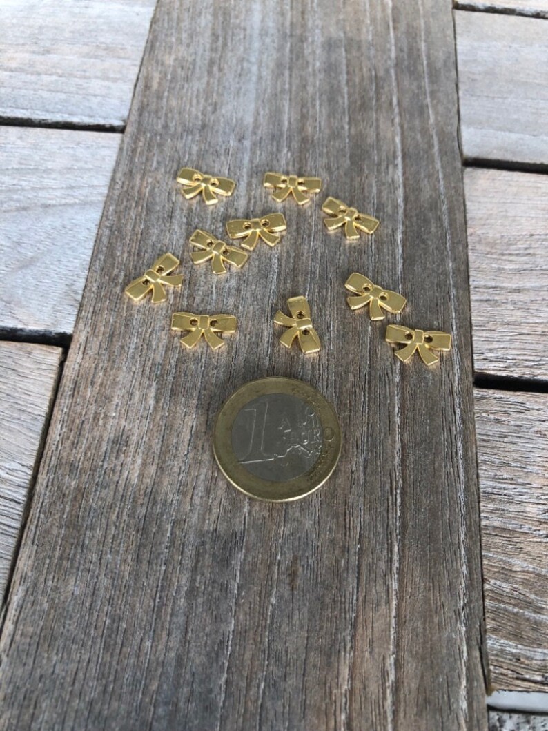 May include: A collection of ten gold-colored metal bow-shaped embellishments scattered on a wooden surface. A euro coin is included for scale.