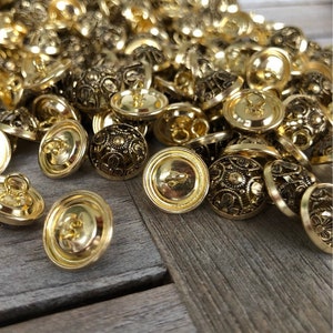 May include: A pile of gold-toned buttons with intricate designs. The buttons are round and have a raised center with a floral pattern. They have a shank on the back for sewing.