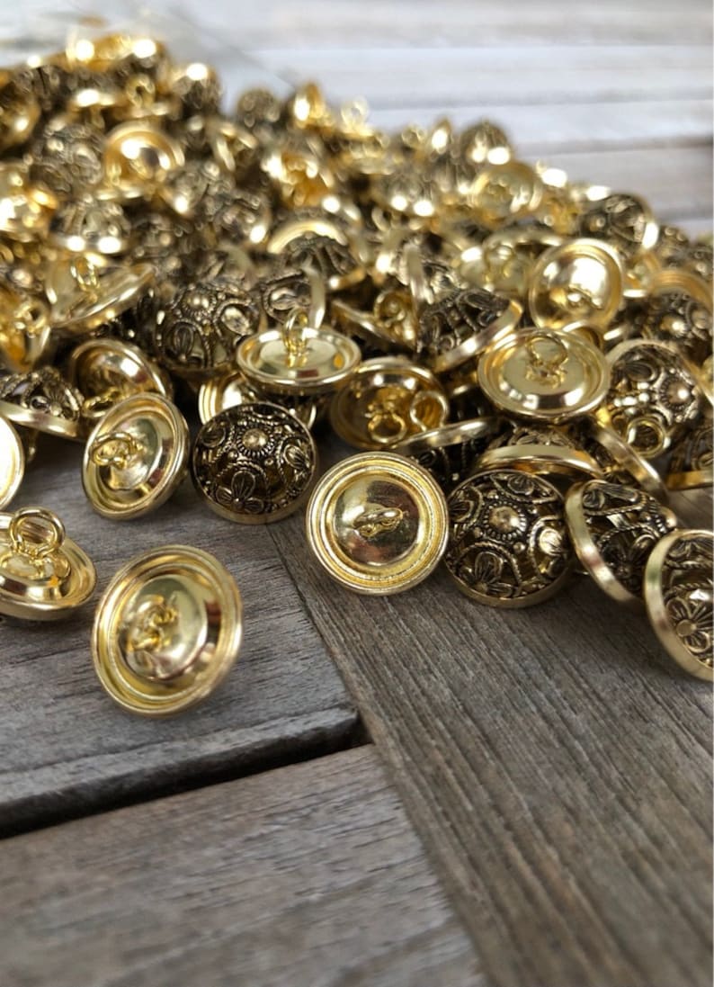 May include: A pile of gold-toned buttons with intricate designs. The buttons are round and have a raised center with a floral pattern. They have a shank on the back for sewing.