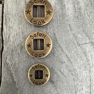 Könnte beinhalten: Drei goldfarbene Metallknöpfe mit der Aufschrift "Safari Look" und einem Stern auf jeder Seite. Die Knöpfe sind in verschiedenen Größen.